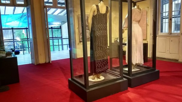 Vista de uma sala com carpete vermelho, e 2 vestidos de &eacute;poca dentro de caixas de vidro em exposi&ccedil;&atilde;o.