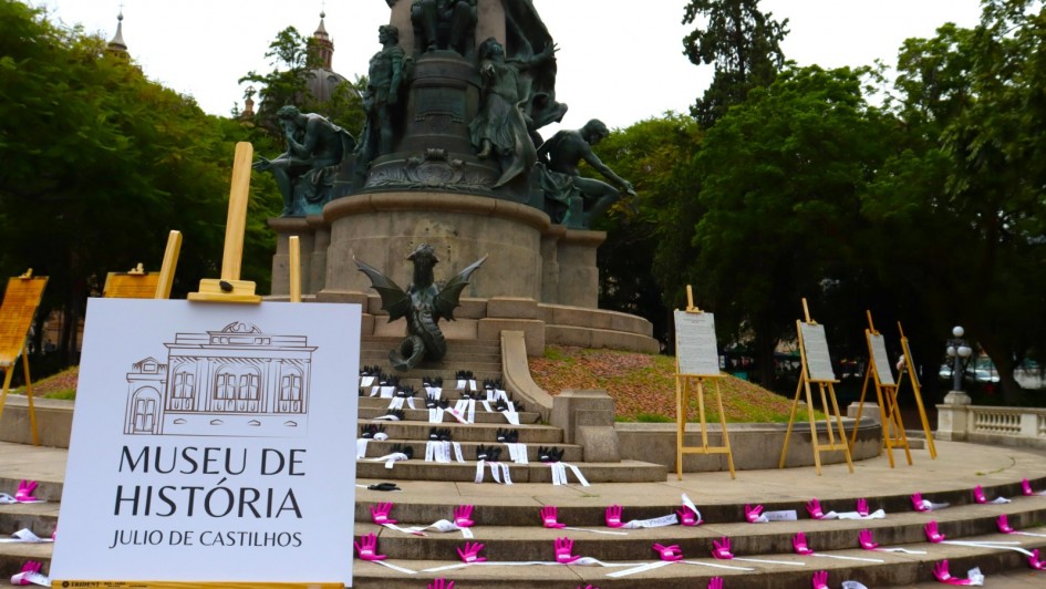 Monumento a Julio de Castilhos na Pra&ccedil;a da Matriz, cercado por &aacute;rvores densas, exposi&ccedil;&atilde;o cultural ao ar livre em frente ao monumento, cavaletes de madeira com pain&eacute;is informativos espalhados pela cena, placa branca em primeiro plano com identidade visual de museu, escadaria de pedra decorada com v&aacute;rias m&atilde;os rosas e faixas brancas distribu&iacute;das artisticamente