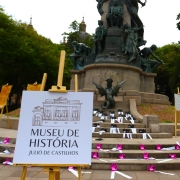 Monumento a Julio de Castilhos na Pra&ccedil;a da Matriz, cercado por &aacute;rvores densas, exposi&ccedil;&atilde;o cultural ao ar livre em frente ao monumento, cavaletes de madeira com pain&eacute;is informativos espalhados pela cena, placa branca em primeiro plano com identidade visual de museu, escadaria de pedra decorada com v&aacute;rias m&atilde;os rosas e faixas brancas distribu&iacute;das artisticamente