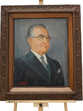 Quadro emoldurado sobre cavalete de madeira com o retrato de Get&iacute;lio Vargas, de terno escuro e &oacute;culos
